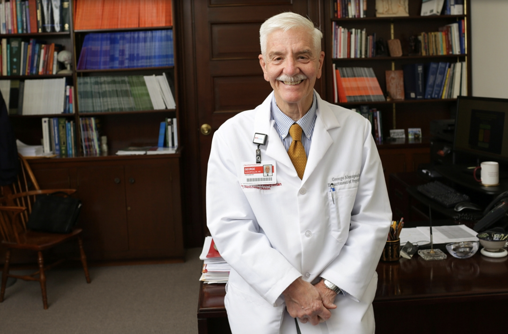 Image of Dr. George Alexopolous smiling and sitting at a desk with books on the bookshelf behind him